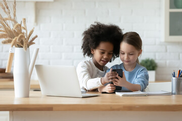 Happy African American and Caucasian little girls using phone together, sitting at table in...