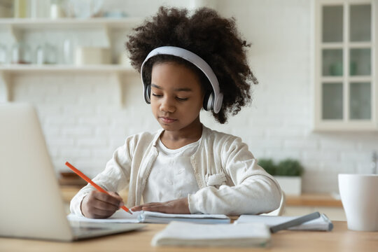 Pretty African American little girl wearing headphones studying at home, sitting at table in modern kitchen, clever child schoolgirl using laptop, listening to lecture, writing notes, homeschooling