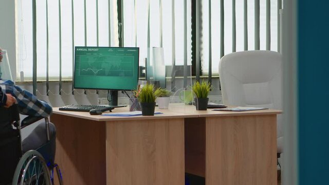 Woman entrepreneur bringing immobilized businessman at workplace with wheelchair wearing protection visor working in new normal business office. Businesspeople with masks respecting social distance