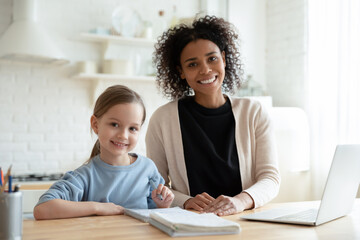 Head shot portrait smiling African American mother with Caucasian daughter studying online, happy mum and adopted child sitting at desk with laptop at home, teacher and pupil doing homework