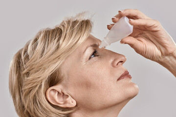 Close up portrait of attractive middle aged woman applying eye drops, standing isolated over grey...