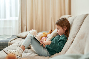 A small cute girl is into a process of playing games on a tablet while sitting on a sofa