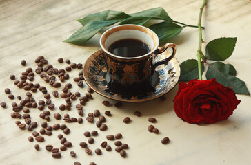 Vintage beautiful cup of hot coffee, bright red rose, green leaves, scattered coffee beans and dark chocolate bar on a wooden table.