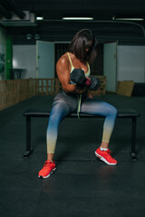 Fototapeta na wymiar Fit young girl with light brown hair wearing black leggins, pink top and sneakers and doing squats with barbell, dark gym at background, cross training workout, portrait.
