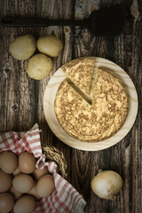 Aerial view of a potato omelette on a wooden board with ingredients: Eggs in a wicker basket, potatoes and onion