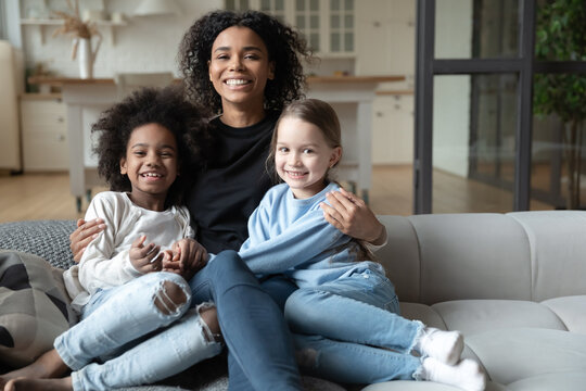 Portrait of smiling African American mother hugging two little daughters, multiracial family concept, sitting on cozy couch in living room, posing for photo, looking at camera, adopted child