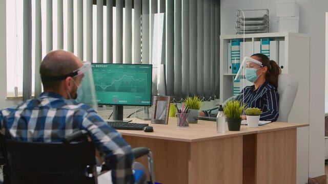 Workers talking wearing protection face masks in new normal office during coronavirus, businessman moving with wheelchair. Immobilized man and coworker in corporate company respecting social distance.