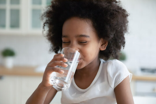 Pretty little African American girl drinking fresh water in kitchen close up, cute preschool child kid holding glass of pure mineral water, enjoying, healthy lifestyle and refreshment concept
