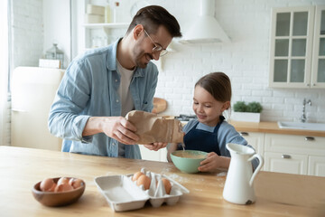 Obraz na płótnie Canvas Smiling father and adorable little daughter cooking dough for pancakes in kitchen, standing at wooden table, happy dad holding flour, helping to cute girl child, family enjoying leisure time together
