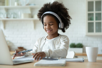 Smiling African American little girl wearing headphones studying at home, sitting at table with...