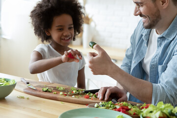 Close up happy young father and adorable African American little daughter playing with vegetables, sitting at wooden table in kitchen, dad and child having fun, multiracial family enjoying weekend