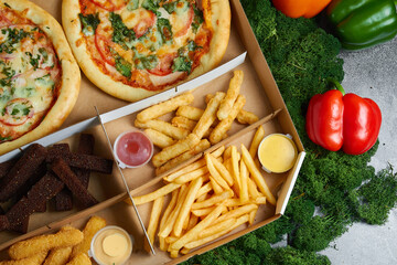 hot snacks, pizza, toasts Borodino, squid rings, spicy chicken wings deep-fried fries with soy sauce in cardboard packaging box against the background of green moss and red and yellow peppers
