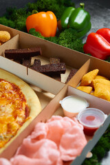 hot snacks, pizza, toasts Borodino, squid rings, spicy chicken wings deep-fried fries with soy sauce in cardboard packaging box against the background of green moss and red and yellow peppers
