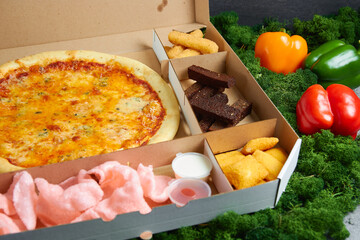 hot snacks, pizza, toasts Borodino, squid rings, spicy chicken wings deep-fried fries with soy sauce in cardboard packaging box against the background of green moss and red and yellow peppers
