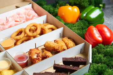 hot appetizers, beard toast, squid rings, spicy chicken wings deep-fried fries with soy sauce in cardboard packaging box against the background of green moss and red and yellow peppers