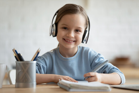 Head shot portrait smiling little girl wearing headphones looking at camera, sitting at table, studying at home, happy pretty child doing homework, holding pen, writing notes, watching webinar