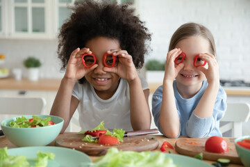 Close up head shot smiling diverse little girls holding red pepper as glasses, sitting at table...