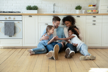 Happy multiracial family with children sitting on warm wooden floor in kitchen, enjoying leisure...