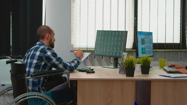 Handicapped paralysed businessman in wheelchair analysing economy data taking notes making financial expertise in business office discussing with colleague. Immobilized man using modern technology.