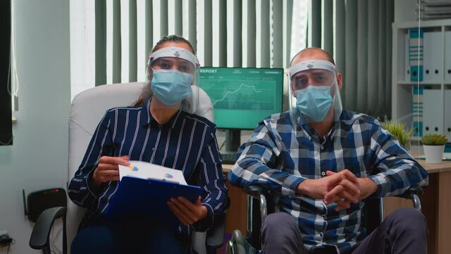 Businessman in wheelchair and coworker wearing visor having videomeeting in business office during global pandemic. Immobilized freelancer working in financial company respecting social distance.
