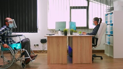 Man with disability and woman with protective face masks keeping distance to prevent covid virus in company working in office with new normal. Coworkers using sanitize alcohol gel against corona virus