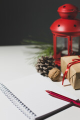 An open notebook and a red pen are on the table and a christmas decoration. Gift with, candlestick, red ribbon, cedar cones and a branch of a coniferous tree. The eve of the holiday. Copy space.
