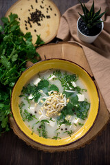 Cold soup broth in a deep yellow bowl with bean sprouts, shredded herbs, feta chunks, black peppercorns. On a wooden round board with a bunch of cilantro leaves and a beige napkin. View from above.