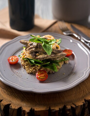 Beef stroganoff cooked in a creamy sauce, with quinoa, cherry tomatoes, basil leaves on a gray ceramic plate on a wooden board. Low calorie portion. Beige napkin, fork and knife on the side. Top view.