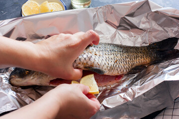 Raw carp with spices and salt on foil before baking, female hands place lemon slices in the belly of the fish, horizontal