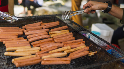 Grilling full of raw fresh sausage on barbecue grill and turned it using tongs on a summer picnic. Preparation of roasted chicken hot dogs on a hot grill stove. Selective focus.
