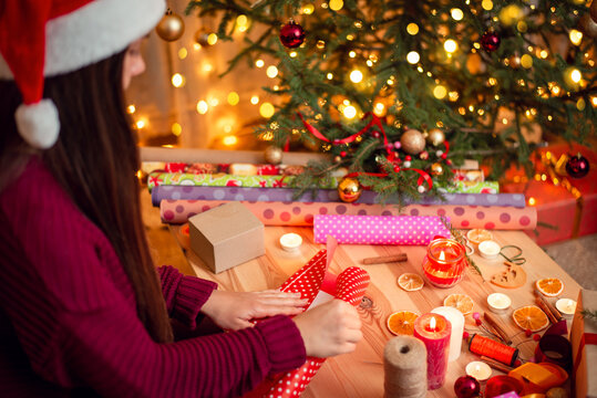 Side view of a girl in santa hat preparing presents to present them to friends in Christmas night. Attractive woman packing boxes into paper near decorated christmas tree