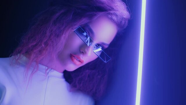 Close-up portrait of charming young woman wearing futuristic glasses and white outfit standing by wall in nightclub and looking into camera. Cyberpunk, female backlit with violet neon light at party