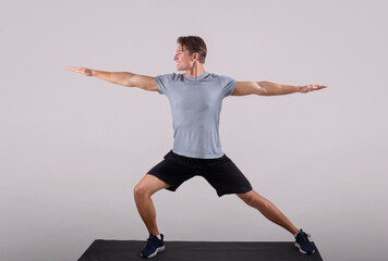 Handsome young man in sportswear doing warrior yoga pose on sports mat over light background, full length portrait - obrazy, fototapety, plakaty