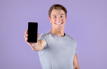 Happy young sportsman showing smartphone with empty screen over lilac background, mockup for mobile app design