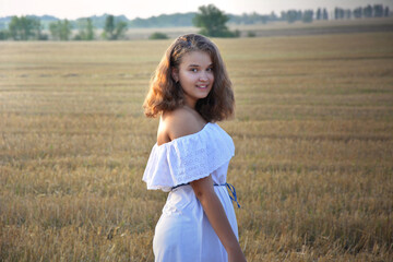 Fototapeta na wymiar A young girl with lush blond hair in a white dress stands against a mown field and smiles