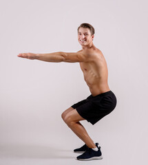 Full length portrait of sexy shirtless guy doing squats, looking at camera and smiling on light...