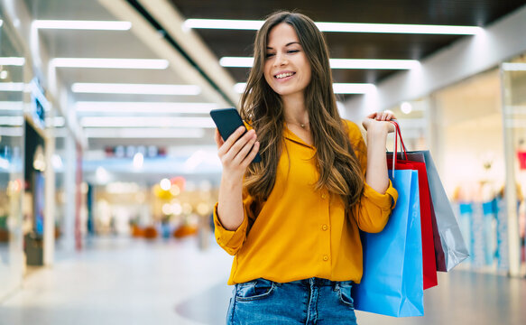 Happy beautiful young stylish woman with shopping bags is using smart phone while walking in the mall on black friday