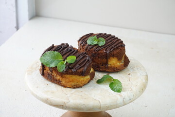 Two chocolate cheesy cake brownie topped chocolate glaze and mint leaves in marble cake tray in cream grainy background. Selective focus  