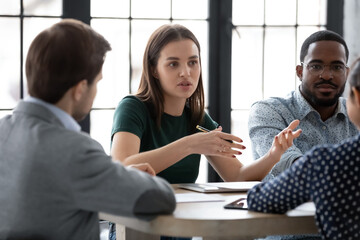 Two diverse multiethnic groups of business partners sitting at negotiation table, concentrated millennial female employee explaining plan project details to interested teammates on meeting in office
