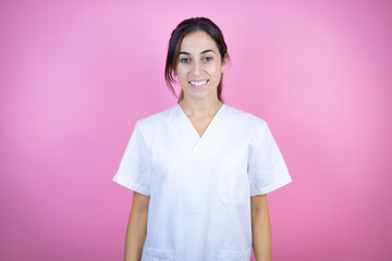 Young brunette doctor girl wearing nurse or surgeon uniform over isolated pink background with a happy face standing and smiling with a confident smile showing teeth