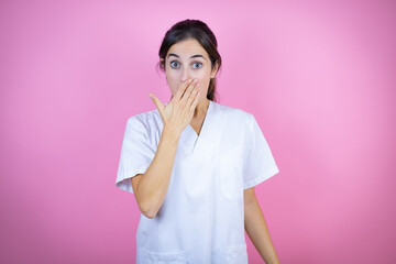 Young brunette doctor girl wearing nurse or surgeon uniform over isolated pink background surprised covering the mouth