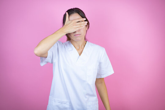 Young brunette doctor girl wearing nurse or surgeon uniform over isolated pink background serious and covering her eyes with her hand