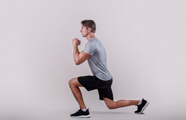 Side view of young fit guy in sportswear doing lunge on light studio background - obrazy, fototapety, plakaty