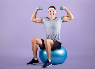 Obraz premium Happy young sportsman sitting on fitness ball and working out with dumbbells on violet studio background, full length
