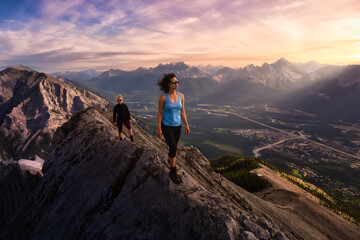 Fototapeta premium Adventurous Girl is hiking up a rocky mountain. Dramatic Colorful Sunset Sky. Taken from Mt Lady MacDonald, Canmore, Alberta, Canada. Epic Adventure Travel