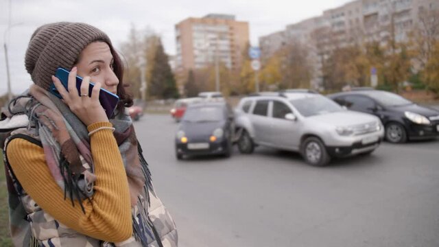 young girl in a hat gesturing emotionally talking on the phone against the background of a car accident. upset looks at the wrecked car and shouts into the phone.