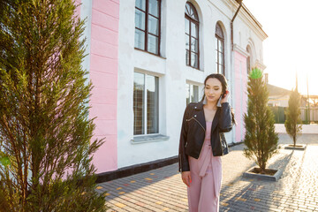 Pretty long-haired young woman in black leather jacket posing on the street by sunset light,...