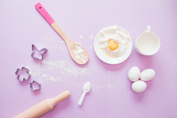 Fototapeta na wymiar Baking ingredients. Eggs, flour, milk, whisk, rolling pin, bakeware on a lilac background, top view. cook's workplace in the kitchen. beautiful dishes. Copy space. homemade cookies. Cooking background