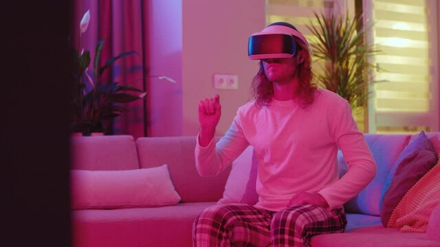 Long-haired caucasian young man staying home in pink neon lights playing VR games online social media apps scrolling web pages using futuristic devices.
