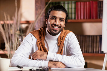 Obraz premium Portrait Of Young Indian Teacher Man Smiling Sitting At Workplace